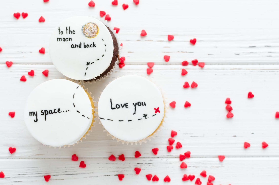 Drei Cupcakes mit weißen essbaren Auflegern und schwarzem Schriftzug, dazu rote Herzstreusel auf weißem Holz